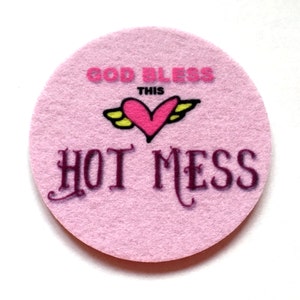 May include: Pink round felt coaster with the text "God Bless This Hot Mess" in black and pink lettering. A pink heart with yellow wings is above the text.