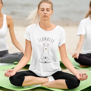 Op de afbeelding: Een vrouw draagt een wit T-shirt met een lama-afbeelding en de tekst "LLAMASTE" en zit in een yogahouding op een groene mat. Ze heeft haar ogen gesloten en mediteert.