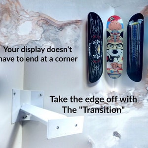 May include: White metal bracket mounted on a wall corner with three skateboards hanging from it. The skateboards are black with graphic designs. The text "Your display doesn't have to end at a corner" and "Take the edge off with The "Transition"" is visible in the image.