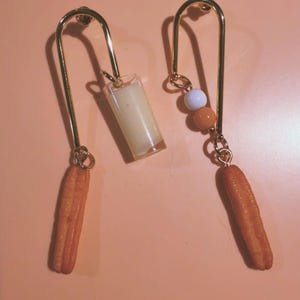 May include: A pair of quirky earrings with a gold-coloured metal design. One earring features a small glass of a creamy liquid, while the other has a cluster of beads and a churro-shaped charm. The background is a solid peach colour.