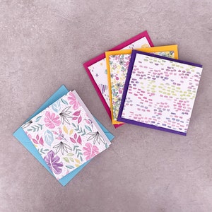 May include: Four greeting cards with different floral and abstract patterns. The cards are stacked on top of each other and have a light blue, yellow, pink, and purple border.