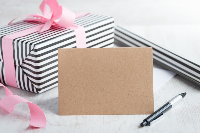 May include: A blank brown card with a black and white striped gift box with a pink ribbon in the background. A black pen is lying on the table.