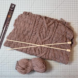 May include: A close-up of knitting supplies including a brown knitted scarf, two wooden knitting needles, a ball of brown yarn, and a clear plastic ruler. The scarf has a cable knit design. The knitting needles are labeled with the size.