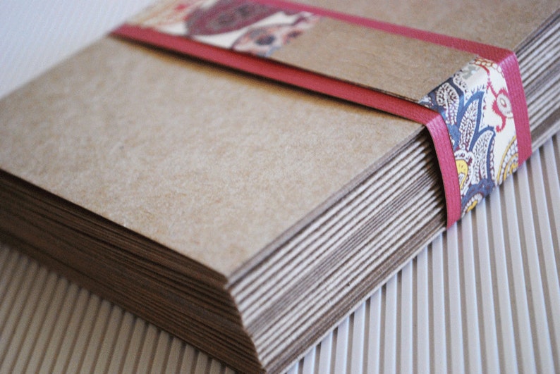 May include: A stack of brown paper cards tied together with a red ribbon and a patterned fabric. The ribbon has a paisley design in blue, white, and yellow.