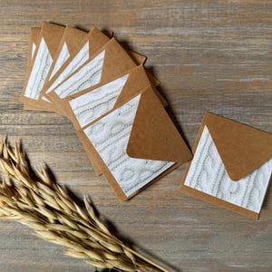 May include: A collection of small, brown paper envelopes with white and gray cable knit pattern inserts. The envelopes are arranged on a wooden surface, with a bundle of dried wheat stalks in the foreground.