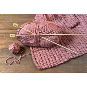Puede incluir: Una imagen de naturaleza muerta de suministros para tejer. Una madeja de hilo rosa es atravesada por dos agujas de tejer de madera. También hay una madeja de hilo más pequeña y una muestra tejida. Las agujas están etiquetadas con los tamaños 3 US y 4 US.