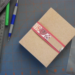 May include: A brown cardboard box with a red and white patterned ribbon. The box is sitting on a gray cutting mat.