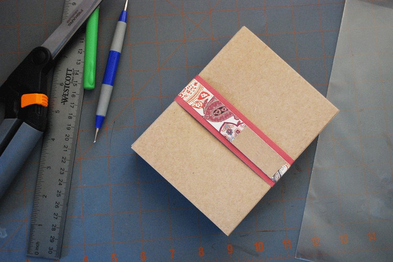 May include: A brown cardboard box with a red and white patterned ribbon. The box is sitting on a gray cutting mat.