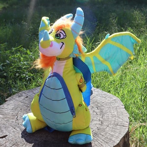 May include: A yellow and blue plush dragon backpack with orange hair and a blue zipper. The dragon is sitting on a tree stump with green grass in the background.
