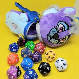May include: A purple and blue plush toy with a zippered pouch, containing a set of colorful dice. The dice are various shapes and sizes, with numbers printed on each side.