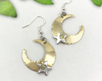 May include: A pair of gold crescent moon earrings with a silver star charm. The earrings have a hammered texture and are suspended from silver hooks.