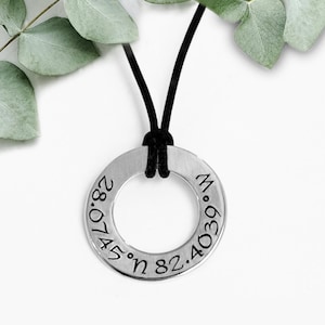 May include: A silver-colored pendant necklace with a black cord. The pendant is a circular ring engraved with coordinates. The necklace is displayed on a white background with green leaves.