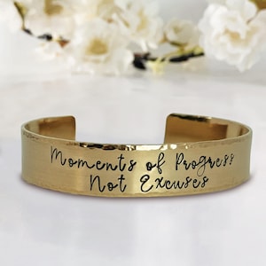 May include: Gold-toned hammered metal bracelet with the inscription "Moments of Progress Not Excuses" in black lettering.