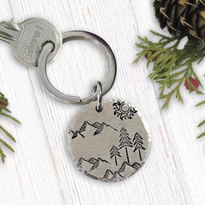 May include: A silver keychain with a round charm featuring a hand-drawn design of mountains, trees, and a sun. The charm is attached to a silver key ring.
