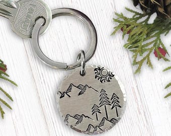May include: A silver keychain with a round charm featuring a hand-drawn design of mountains, trees, and a sun. The charm is attached to a silver key ring.