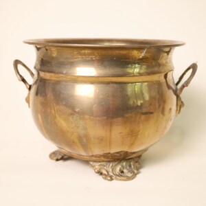 Puede incluir: Una jardinera de latón vintage con dos asas y patas ornamentadas. La jardinera redonda tiene un borde ancho y un acabado pulido, que refleja la luz. El latón tiene un tono dorado cálido, lo que sugiere edad y uso. Una pieza decorativa para la decoración del hogar.