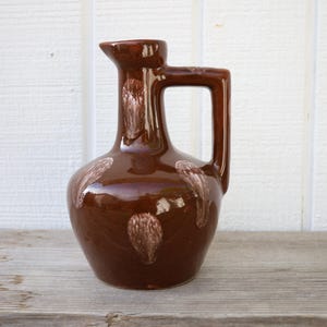 May include: A brown ceramic pitcher with a white design. The pitcher has a wide base and a narrow neck with a handle.