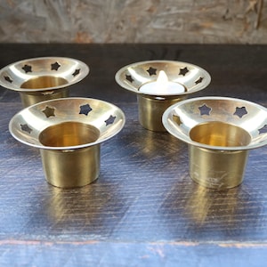May include: Four gold metal candle holders with star-shaped cutouts. The candle holders are arranged in a square shape on a dark wooden surface. One of the candle holders has a white tea light candle inside.