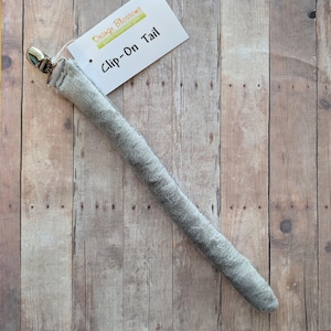 May include: A gray clip-on tail made of soft, plush material. The tail has a silver-colored metal clip at the top for easy attachment. The tag reads "Clip-On Tail" and has the "Orange Blossoms" logo.