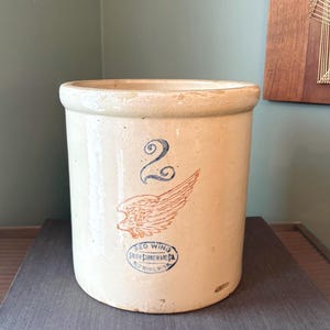 Könnte beinhalten: Ein Vintage-Steingutkrug mit beigefarbenem Finish. Er hat eine blaue Zahl 2 und ein rotes Flügeldesign. Die Worte "RED WING" und "STONEWARE CO." sind auf dem Krug aufgedruckt.