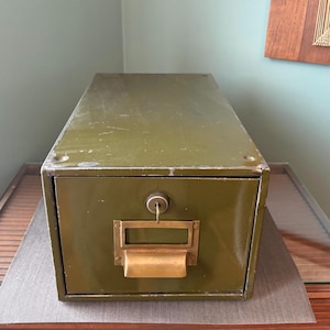 May include: An olive green metal file cabinet with a brass handle and keyhole. The cabinet is rectangular and sits on a wooden surface. The cabinet has a label holder and a key in the lock.