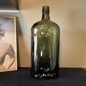 May include: A dark green glass bottle with a textured surface and a round, indented design. The bottle has a wide mouth and a short neck.