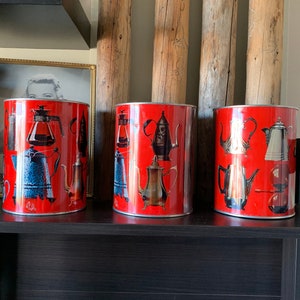 Puede incluir: Tres botes de metal rojo con un diseño vintage que presenta imágenes de cafeteras y teteras. Los botes están en un estante negro.