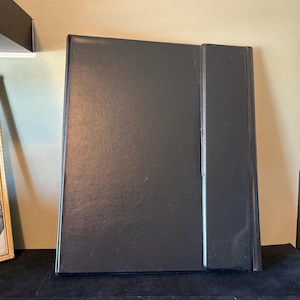 May include: A black album with a raised spine, standing upright. The album is on a dark surface. A framed black and white photograph is visible to the left of the album.