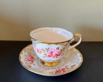 May include: A white teacup and saucer with a pink rose pattern and gold trim. The cup has a delicate handle and the saucer has a scalloped edge.
