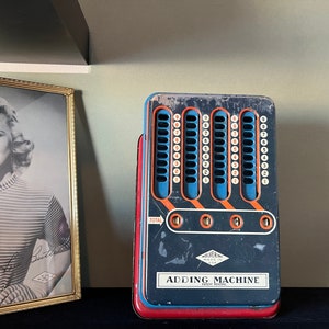 Op de afbeelding: Vintage rekenmachine met een blauw, rood en oranje kleurenschema. De machine heeft meerdere kolommen met nummerknoppen en de tekst "ADDING MACHINE" is zichtbaar. Een ingelijste zwart-witfoto staat op de achtergrond.