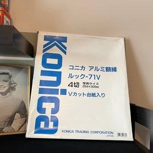 May include: A white box with blue lettering that reads "Konica Aluminium Frame Look-71V 4-cut Photo Size 254x305mm V-Cut Mount Included Konica Trading Corporation Japan".