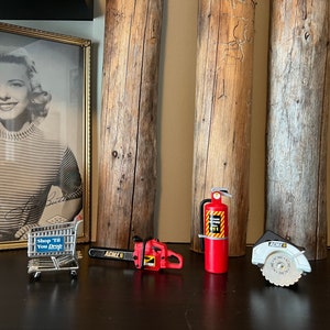 May include: Close-up view of miniature toy tools and equipment displayed on a dark surface against a backdrop of aged wooden planks and a framed black and white photo of a woman. The miniature items include a shopping cart with the text 'Shop 'Til You Drop', a chainsaw labeled 'Agile 8', a red fire extinguisher, and a circular saw. The photo shows a woman in a striped shirt. The overall theme is a collection of miniature tools and equipment.