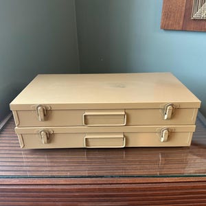 May include: Two stacked, tan metal storage cases with silver clasps and handles. The cases are rectangular and sit on a glass-topped wooden cabinet. The background is a muted green wall.