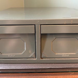 May include: An olive-green metal file cabinet with two drawers. Each drawer features a rectangular, recessed panel and the word "Oxford" printed at the bottom. The cabinet is on a glass surface.