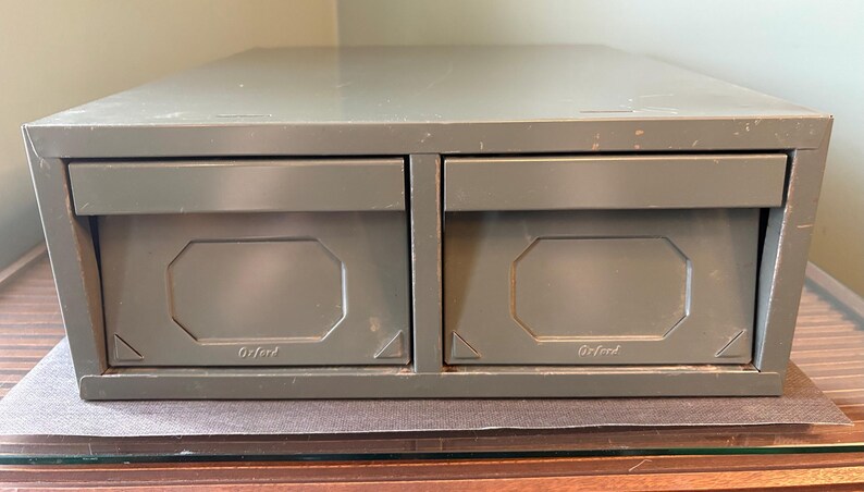 May include: An olive-green metal file cabinet with two drawers. Each drawer features a rectangular, recessed panel and the word "Oxford" printed at the bottom. The cabinet is on a glass surface.