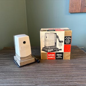 May include: An off-white and silver Boston pencil sharpener with a matching box. The box is tan, red, and black with the words "Boston Vacumette Portable Pencil Sharpener". The sharpener is on a wooden surface.