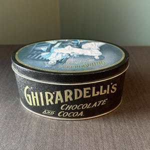 May include: A black Ghirardelli's Chocolate and Cocoa tin with a gold rim and lettering. The lid features a vintage illustration in blue and white. The tin is round and sits on a brown surface.