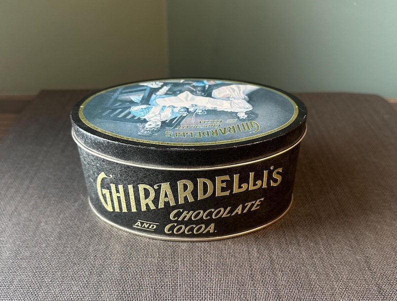 May include: A black Ghirardelli's Chocolate and Cocoa tin with a gold rim and lettering. The lid features a vintage illustration in blue and white. The tin is round and sits on a brown surface.