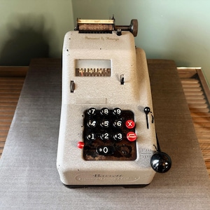Puede incluir: Una antigua máquina de sumar Barrett en color gris claro. La máquina tiene un teclado negro con números blancos y botones rojos "X" y "0". También se ven una manija negra y una pequeña ventana de visualización.