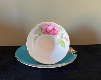 May include: A white teacup and saucer with a pink rose design and a turquoise blue rim. The saucer has a gold trim.