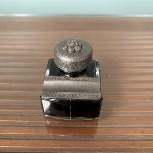 May include: A square black glass inkwell featuring a decorative metallic lid with a raised floral motif and a matching metallic collar. The lid has a textured, ridged edge, and the metal components exhibit a dark, aged finish with intricate detailing.