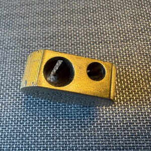 May include: A vintage gold-colored pencil sharpener with two holes of different sizes. The sharpener has a rectangular shape with a ridged top and is slightly worn, suggesting age and use. The object is on a textured surface.