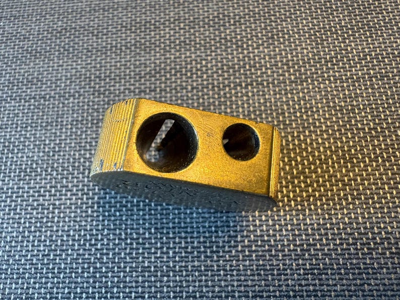 May include: A vintage gold-colored pencil sharpener with two holes of different sizes. The sharpener has a rectangular shape with a ridged top and is slightly worn, suggesting age and use. The object is on a textured surface.