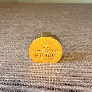 May include: A gold-colored, cylindrical object with the text "50/28", a logo, and "AW FABER MENTOR GERMANY" embossed on its side. The object has a textured, glittery top and a flat, circular base. The background is a textured, gray surface.