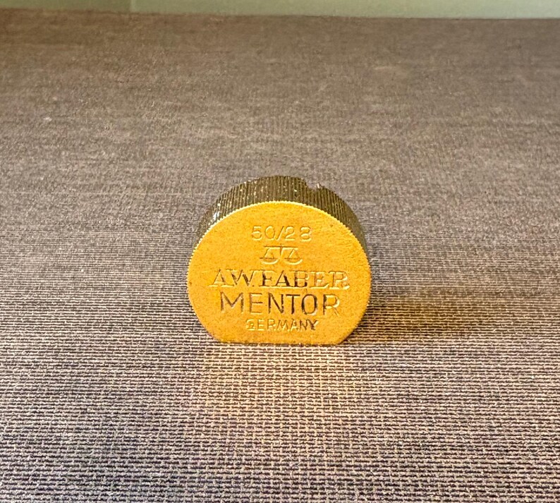 May include: A gold-colored, cylindrical object with the text "50/28", a logo, and "AW FABER MENTOR GERMANY" embossed on its side. The object has a textured, glittery top and a flat, circular base. The background is a textured, gray surface.