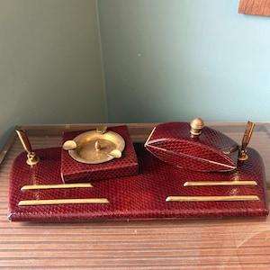 May include: A vintage desk set in a rich burgundy color with a snakeskin pattern. The set includes a pen holder, ashtray, and a small box with a gold-colored knob. The set is placed on a glass surface.