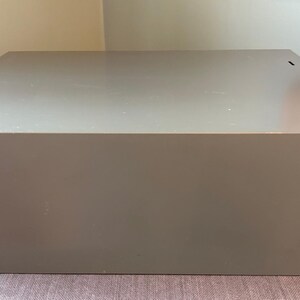 May include: A large, rectangular, grey storage box with a flat top. The box is made of a solid material and has a simple, minimalist design. The box is sitting on a patterned surface.