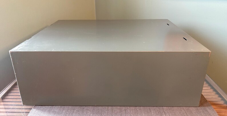 May include: A large, rectangular, grey storage box with a flat top. The box is made of a solid material and has a simple, minimalist design. The box is sitting on a patterned surface.