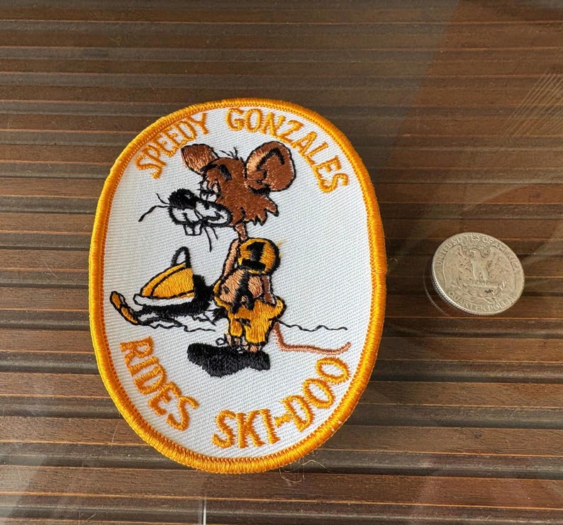 May include: Oval patch featuring Speedy Gonzales riding a ski-doo. The patch has a white background with an orange border and text. The cartoon mouse is brown and yellow. A quarter is next to the patch.