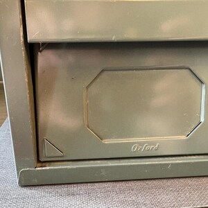 May include: Close-up of a vintage olive green metal file cabinet drawer. The drawer has a recessed panel with the name "Oxford" embossed on it. The cabinet appears to be made of sturdy metal.